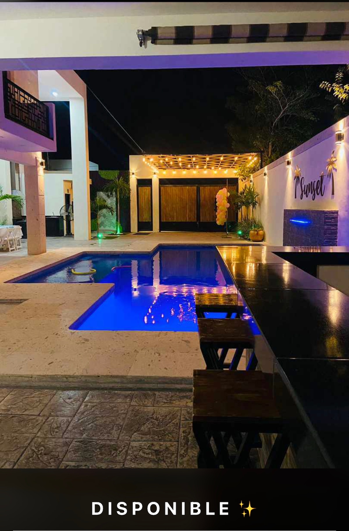 A serene outdoor space is showcased, featuring a pool illuminated with soft blue lighting. A bar with wooden stools lines one side, while elegant overhead lights create a calm ambiance. The area is surrounded by greenery and softly lit pathways, enhancing the tranquil atmosphere.