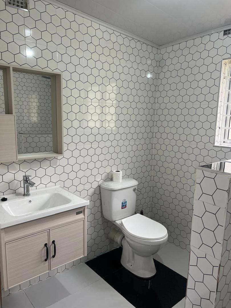 The bathroom features a clean design with hexagonal tiles adorning the walls. A compact vanity with a sink and mirror is positioned next to a toilet. Natural light enters through a window, creating a bright and airy atmosphere.
