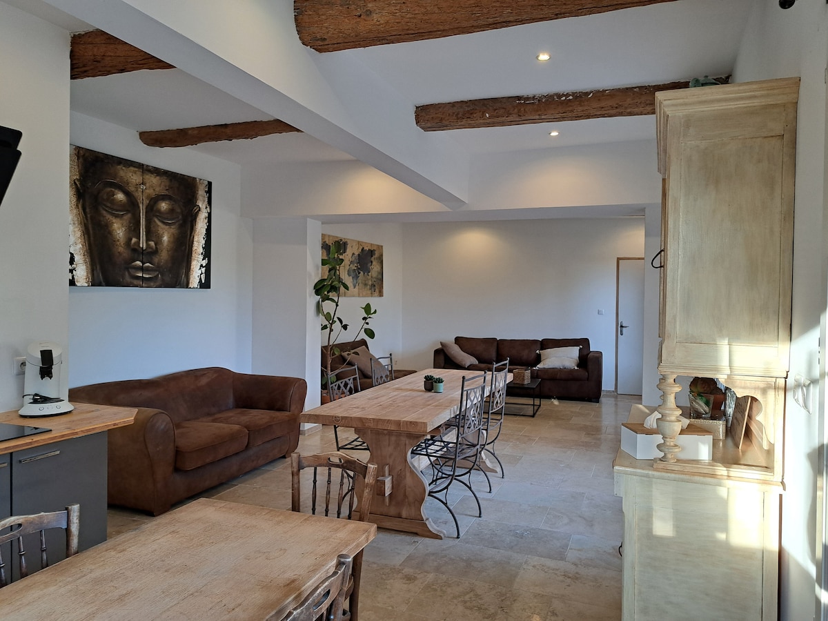A spacious living area features high ceilings with wooden beams and neutral-colored walls. Two comfortable sofas are positioned across from a dining table with wrought-iron chairs. A wooden cabinet is visible, along with modern kitchen appliances, creating an inviting shared space.