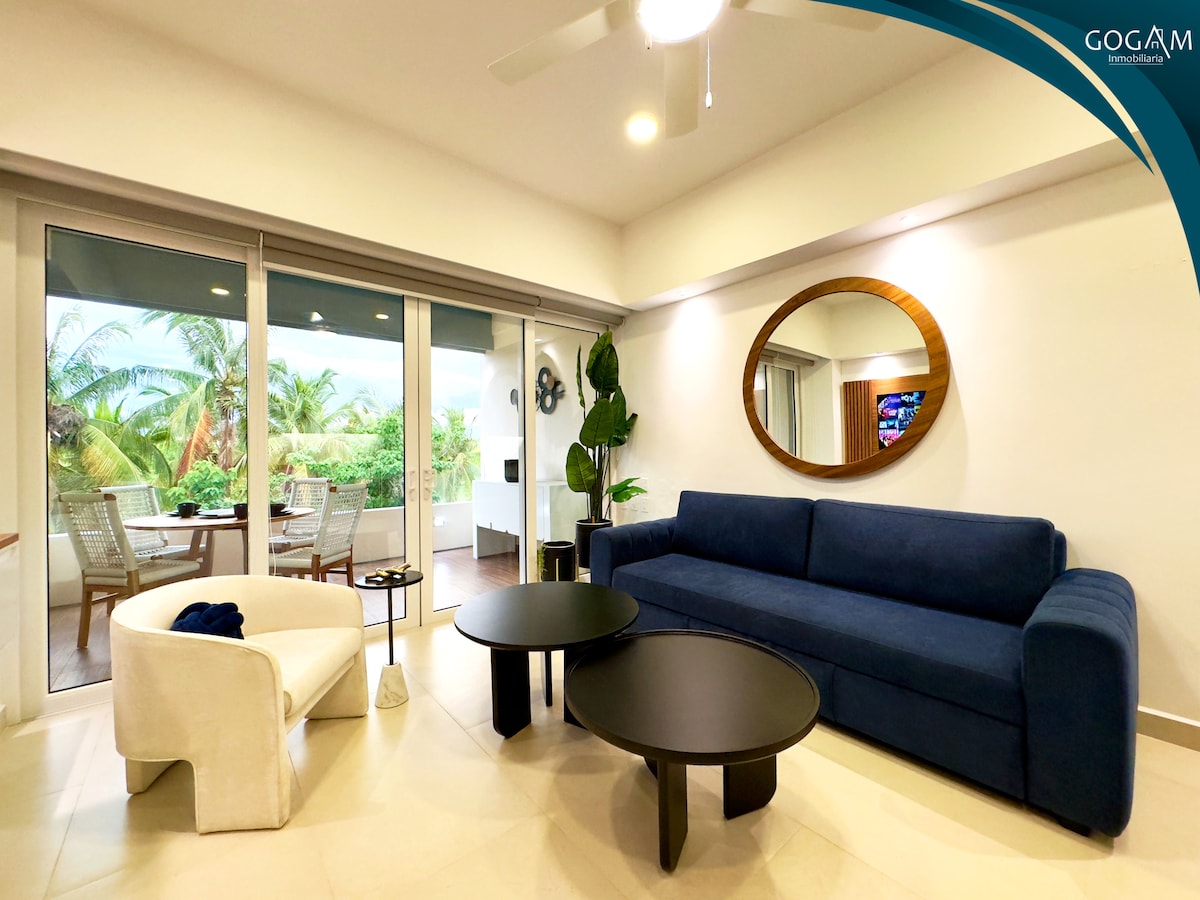 A modern living area features a blue sofa and a white accent chair, complemented by two black coffee tables. Large glass doors provide access to a private balcony with views of lush greenery. A round mirror enhances the spacious feel of the room.