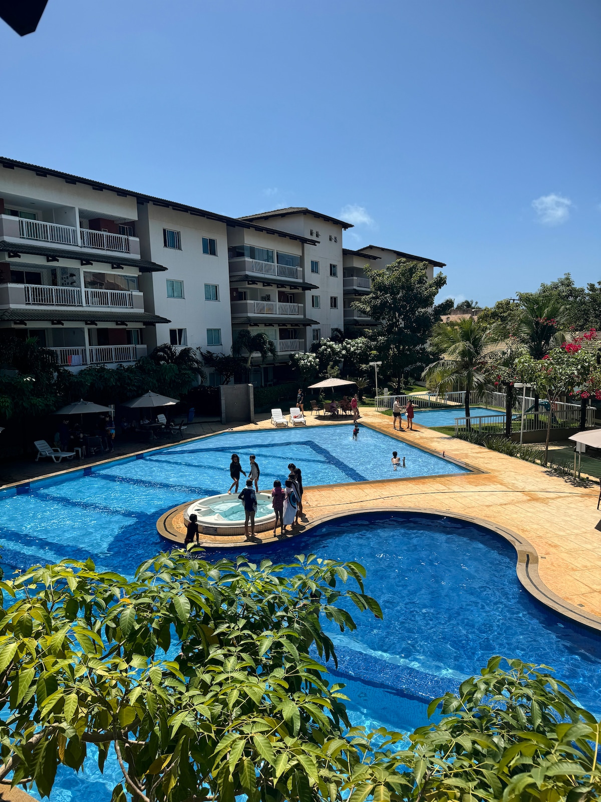 A spacious outdoor pool area is surrounded by well-maintained gardens and palm trees. Various guests can be seen enjoying the water, with sun loungers positioned nearby. The three-story building in the background features balconies and large windows. Clear blue skies complete the relaxing atmosphere.