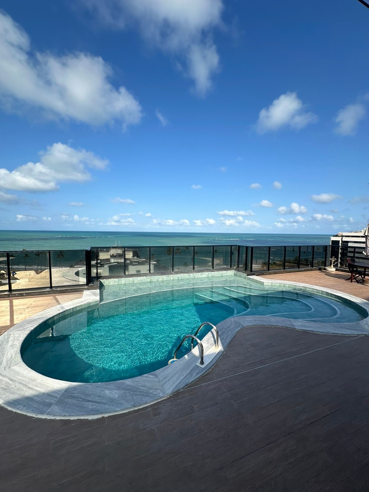 An outdoor pool is positioned by a deck with stunning ocean views. The pool features a unique shape and steps leading into the water, surrounded by glass fencing. A clear blue sky with scattered clouds enhances the atmosphere.