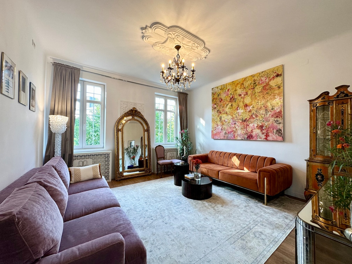 An elegant living room showcases two plush sofas, one in soft purple and the other in warm orange. A large decorative mirror frames a lush indoor plant, while a vibrant wall art piece adds color. Natural light filters through the large windows, highlighting the ornate chandelier.