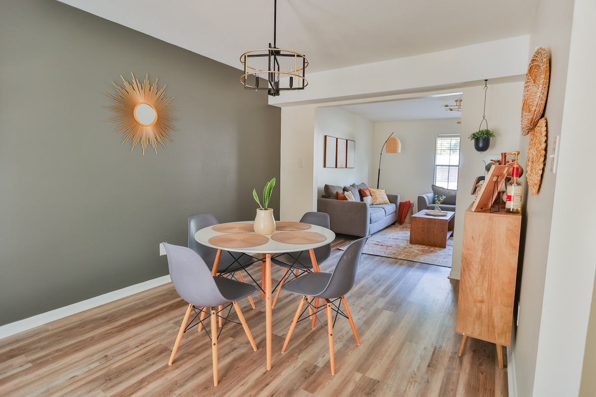 A dining area is featured with a round table surrounded by four modern chairs. A decorative sunburst mirror adorns the wall, complementing the neutral color palette. The open layout leads to a cozy living space visible in the background, where natural light enhances the inviting environment.