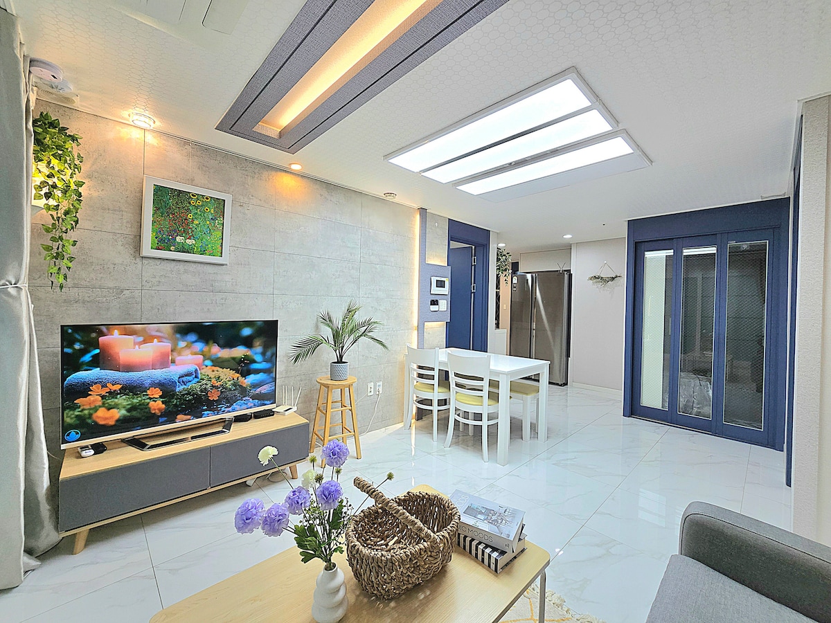 The living area features a large television displaying a tranquil scene, with a modern dining table set for four nearby. Skylights provide ample natural light, and potted plants add greenery to the space. The decor includes neutral tones and minimalist design elements.
