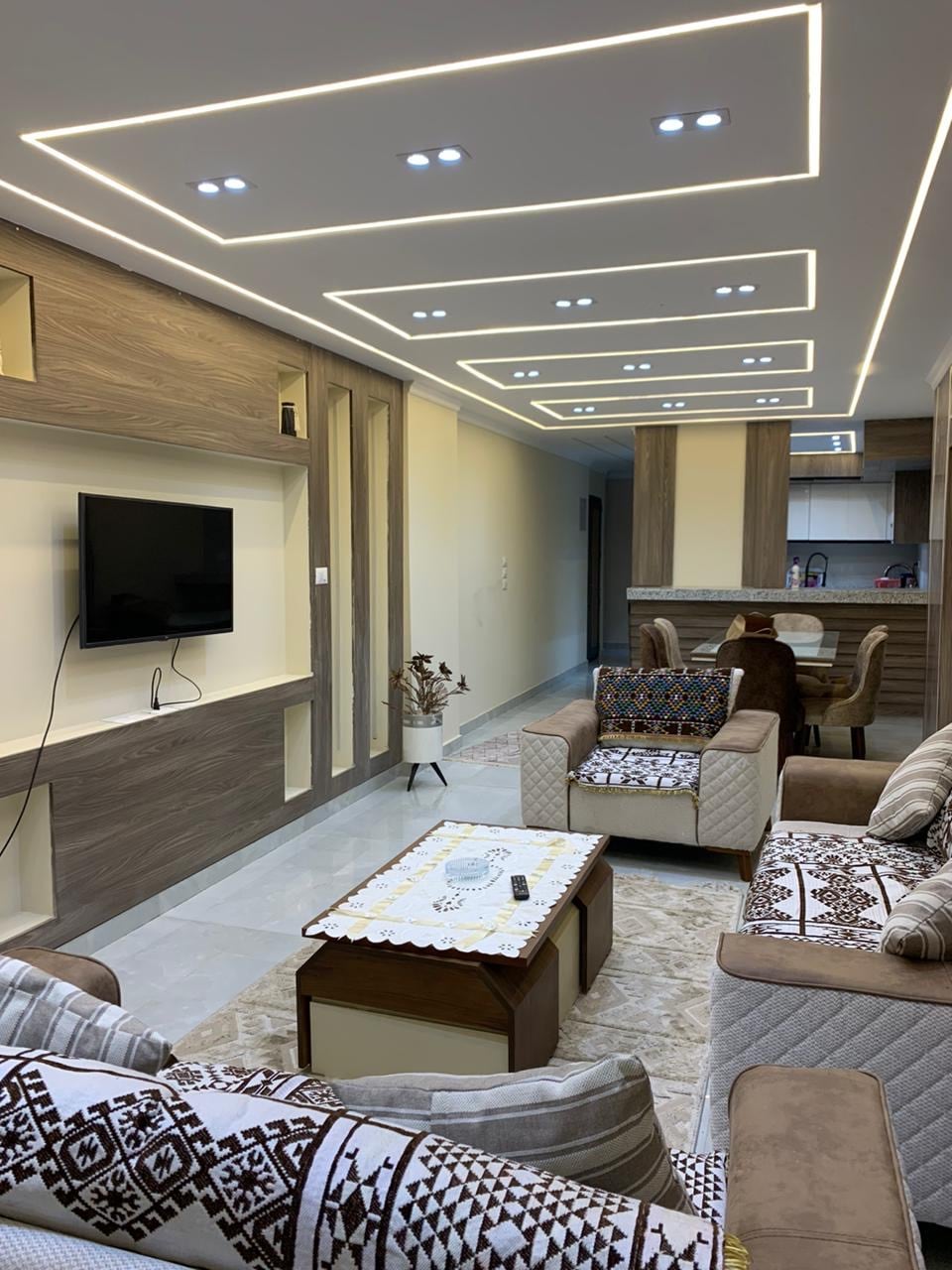A modern living area is showcased, featuring a comfortable seating arrangement with patterned cushions. A wall-mounted television is visible above a stylish wooden console. The ceiling is elegantly designed with backlighting, and a dining area can be seen in the background, enhancing the sense of space.