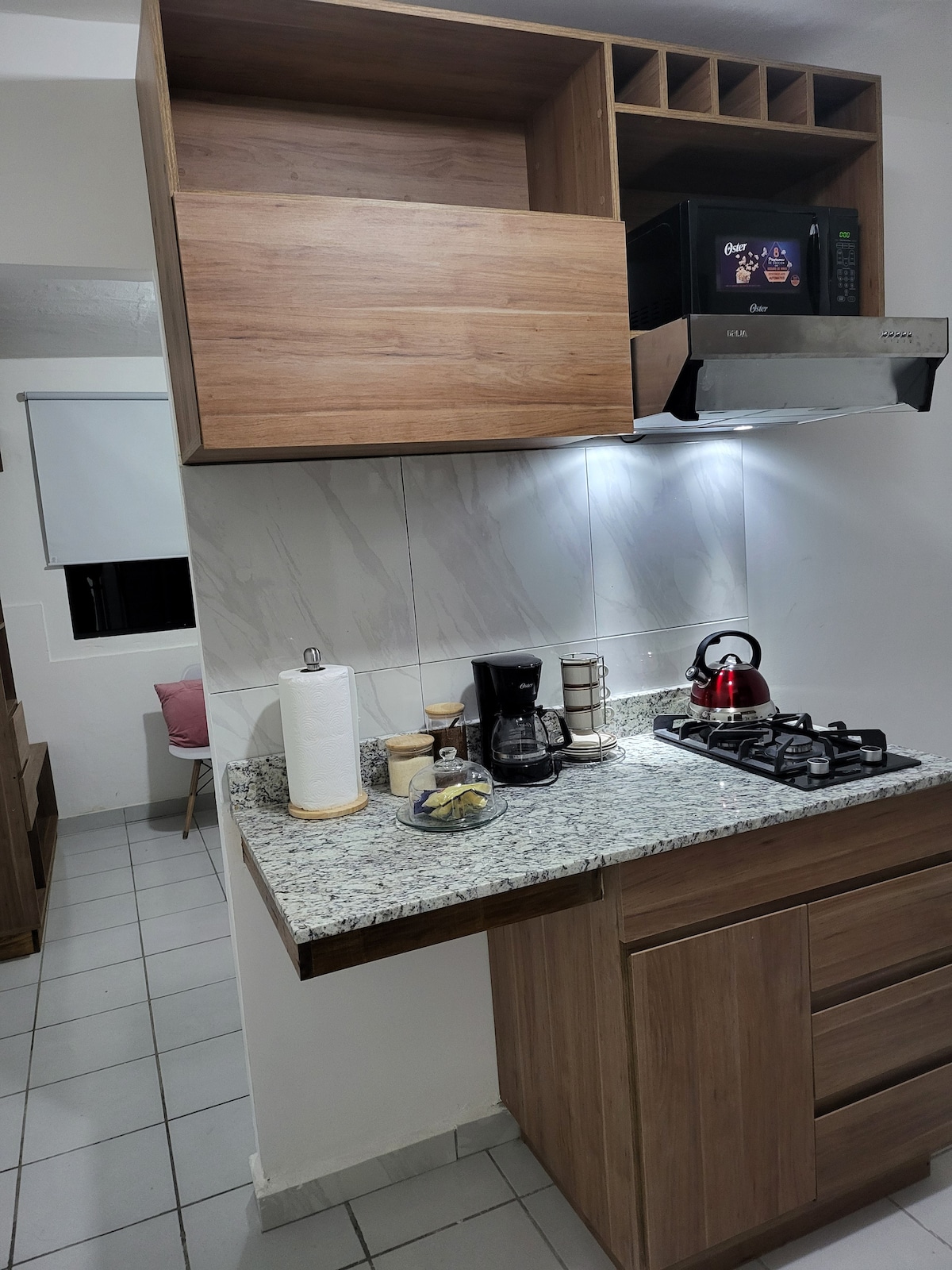 A compact kitchen area features wooden cabinetry and a granite countertop. A coffee maker and kettle are positioned next to a gas stove. Efficient lighting illuminates the space, with a microwave installed above. Storage shelves are incorporated, providing additional organizational options.