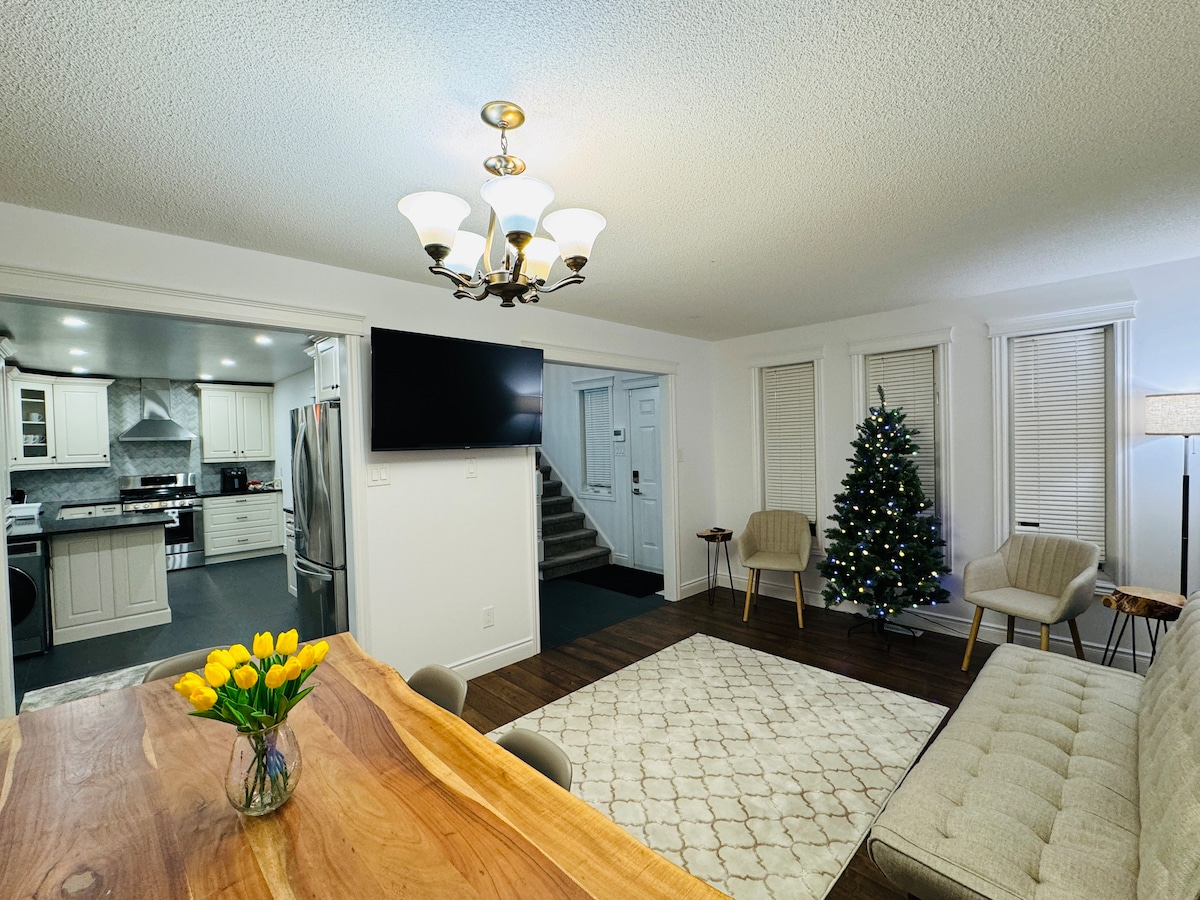 A dining area features a wooden table complemented by a vase of yellow tulips. A cozy seating arrangement includes a light-colored couch and two armchairs. A well-decorated Christmas tree stands nearby, while a wall-mounted television and a chandelier enhance the space's illumination.