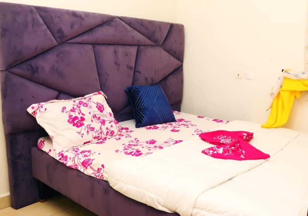 A chic bed is centered in the room, featuring a modern purple headboard with geometric patterns. The bedding displays a floral print in pink tones, complemented by a dark blue decorative pillow. Soft light brightens the space, enhancing the tranquil atmosphere.