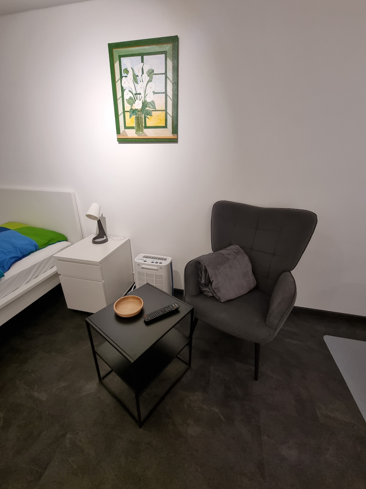 A small seating area is presented with a dark chair and a matching side table. A bowl and remote control are placed on the table. A bedside lamp is positioned beside a bed adorned with a blue and green blanket. A framed artwork displays flowers on a window.