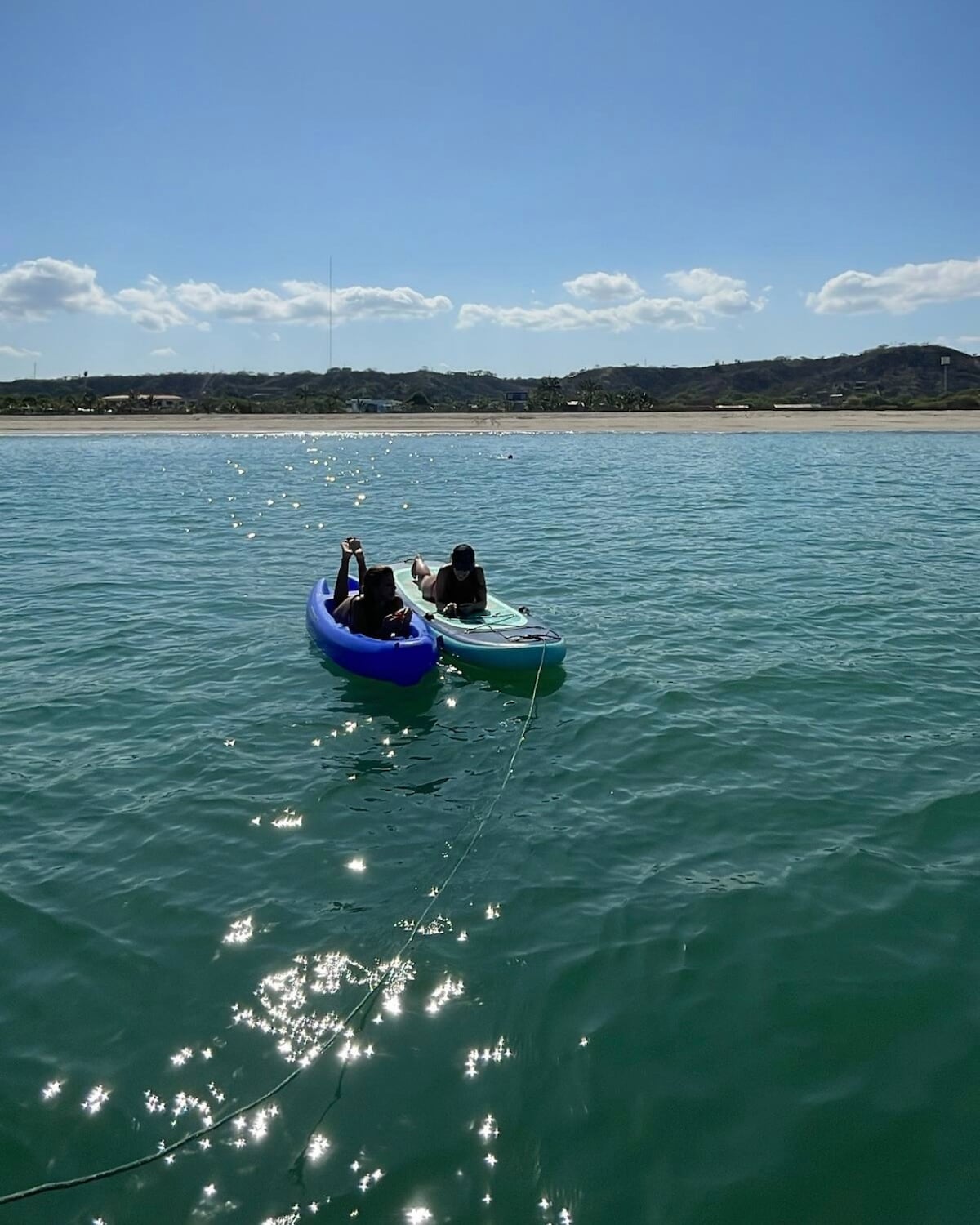 Two kayaks are seen floating on calm turquoise water, with a sandy beach and distant hills visible in the background. A clear sky with a few clouds adds to the serene atmosphere, creating a sense of tranquility on the water.