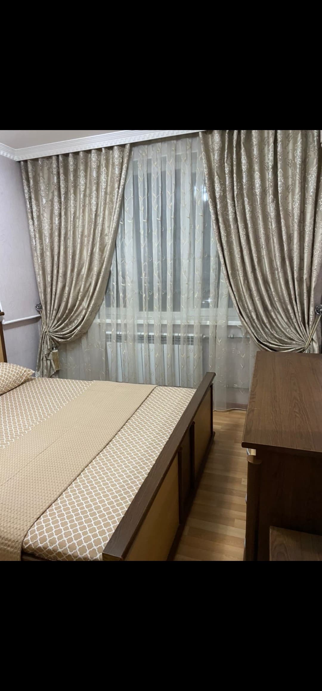 A serene bedroom features a spacious bed covered with a patterned bedspread. Floor-to-ceiling sheer curtains allow natural light to filter through, while rich drapes frame the window. A wooden desk complements the warm tones of the room, enhancing its overall calm ambiance.