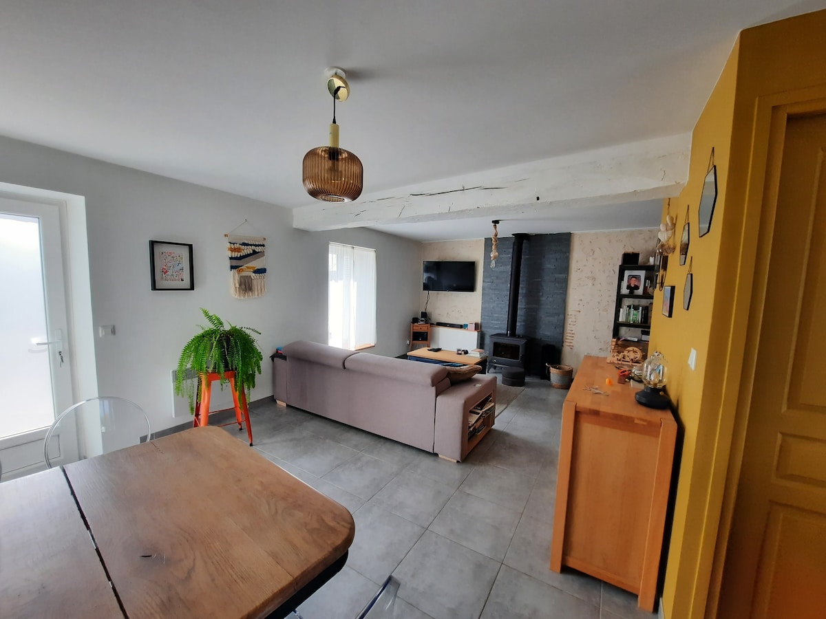 A spacious living area features a combination of light and warm tones. A comfortable sofa is positioned in front of a black wood stove, while a television is mounted on the wall. A large window allows natural light, and an indoor plant adds greenery.
