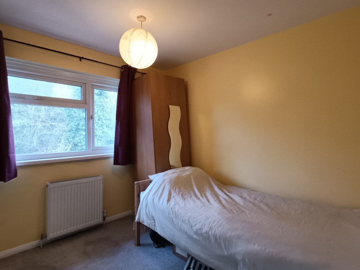 A well-appointed room features a spacious bed covered with a light-colored blanket. A tall wooden wardrobe is positioned beside the bed, with a small nightstand placed nearby. Natural light enters through a large window, framed by burgundy curtains, illuminating the soft yellow walls.
