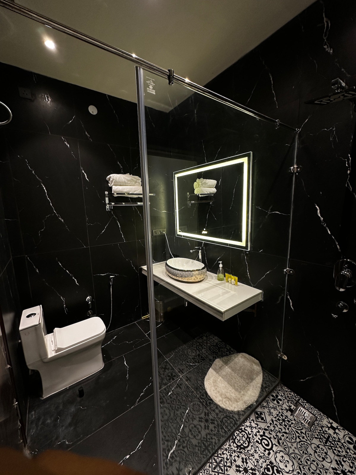 The bathroom features dark marble walls and sleek modern fixtures. A glass shower is prominently displayed alongside a stylish countertop with a round basin. Soft lighting is provided by a mirror with integrated lighting, and decorative tiles adorn the floor.