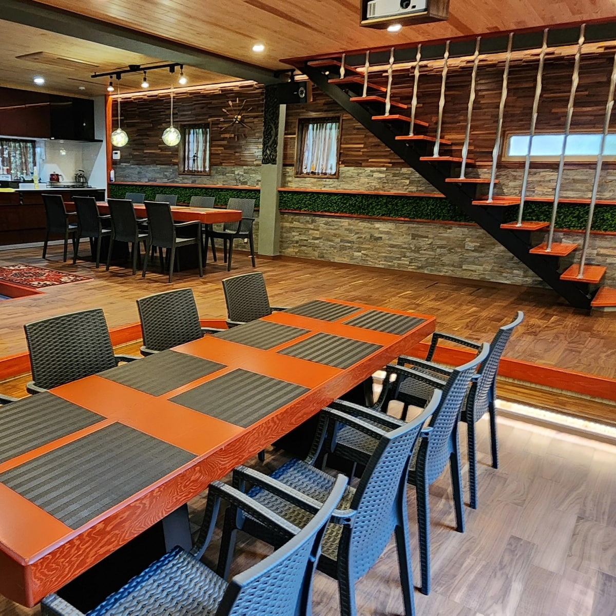 A spacious dining area features a long red table surrounded by six black chairs. Natural wood accents are present in the ceiling and flooring. A staircase with wooden steps and rope railing leads to an upper level, while soft lighting adds a warm touch to the space.