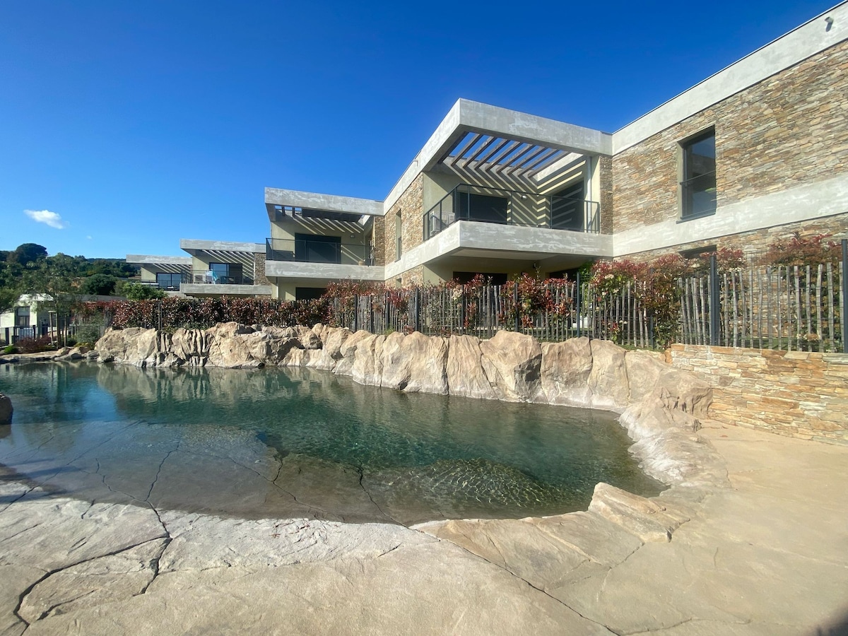 The contemporary building features a combination of stone and modern architectural elements. A rocky shoreline leads to a serene pool nestled among natural landscaping. Lush vegetation frames the structure, enhancing the inviting atmosphere of the outdoor space.
