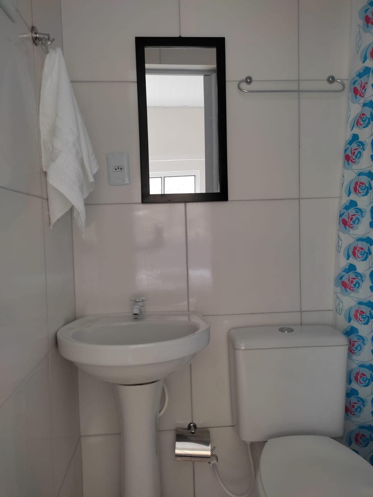 A clean and functional bathroom is presented, featuring a wall-mounted sink with a sleek faucet and a compact toilet. A full-length mirror is mounted above the sink, and a towel is neatly hung nearby. A shower curtain with a floral pattern adds a touch of color to the space.