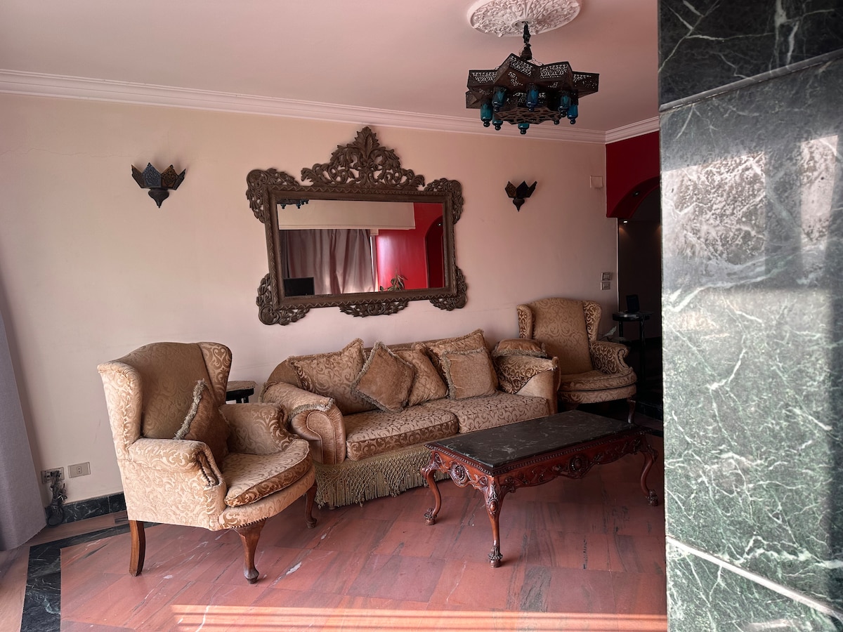 A stylish living area is furnished with an ornate sofa and matching armchairs, complemented by a decorative mirror on the wall. A dark wooden coffee table with intricate detailing occupies the center. Natural light filters in through curtains, creating a warm ambiance.