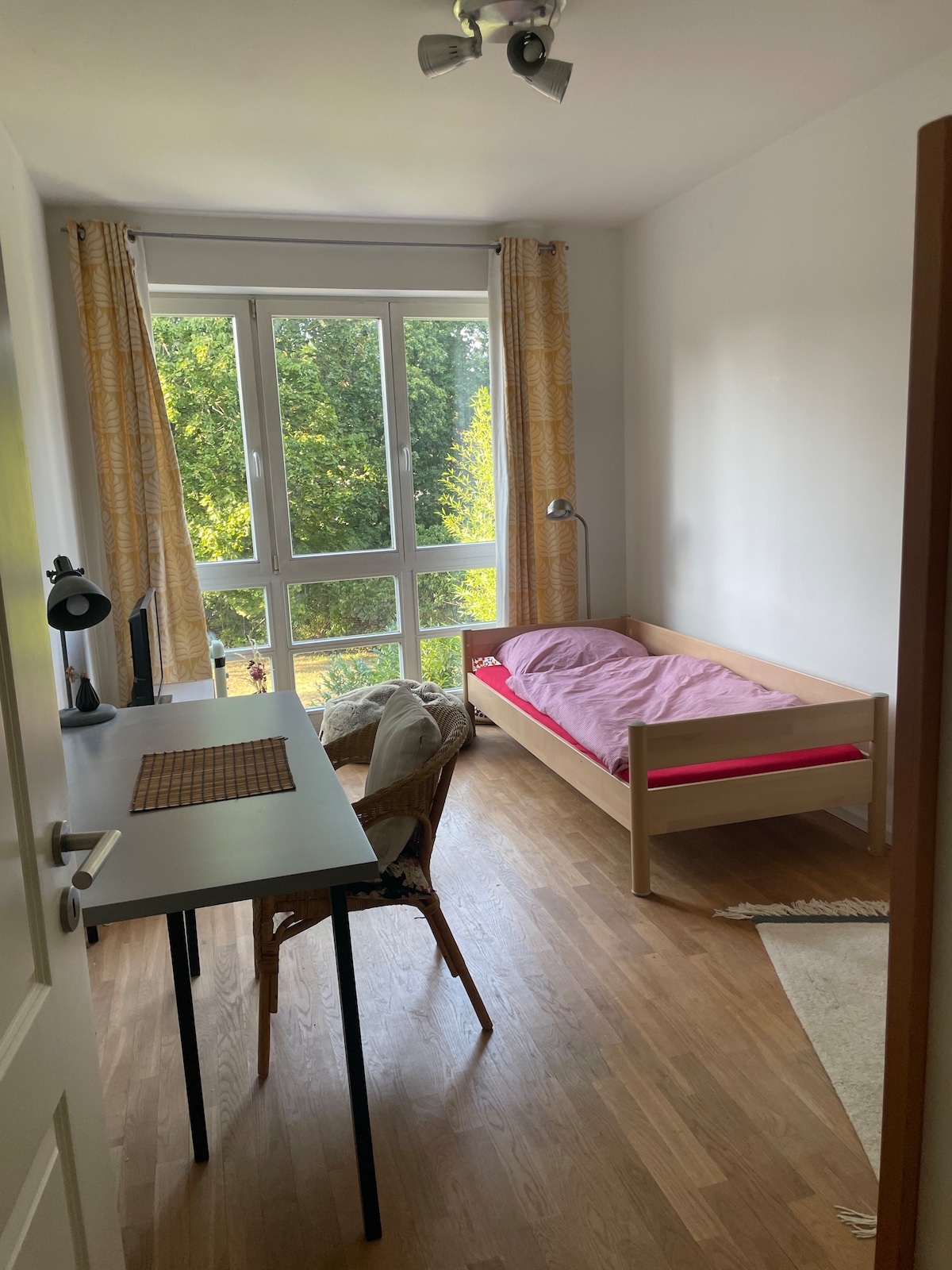 A cozy room is presented, featuring a single bed with pink bedding and a window dressed with light yellow curtains. A desk and chair are situated nearby, alongside a small lamp. Natural light floods the space, highlighting the warm wooden floor.
