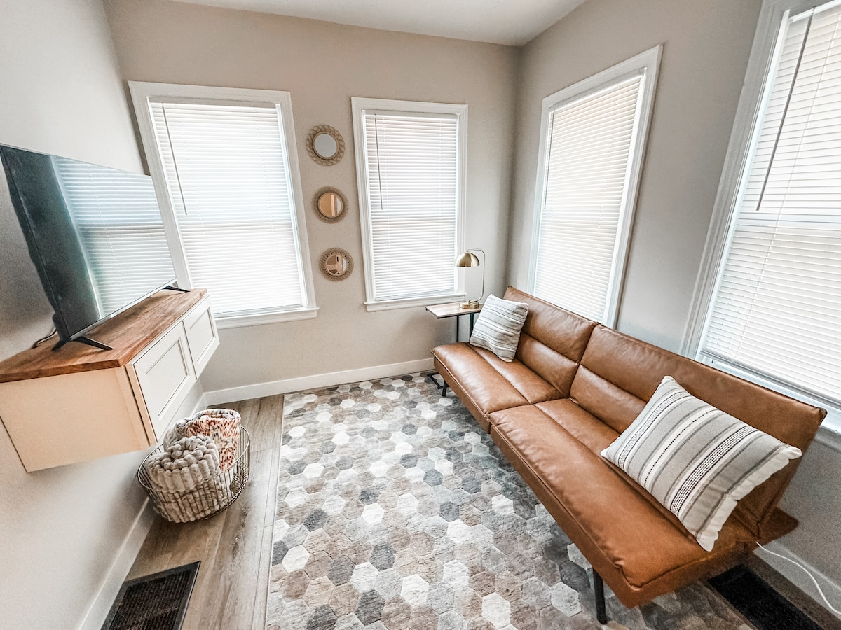 A cozy living area is equipped with a sleek, brown leather futon and a wall-mounted TV. Natural light enters through three windows adorned with blinds. A patterned rug and a side table enhance the space, creating a welcoming environment.