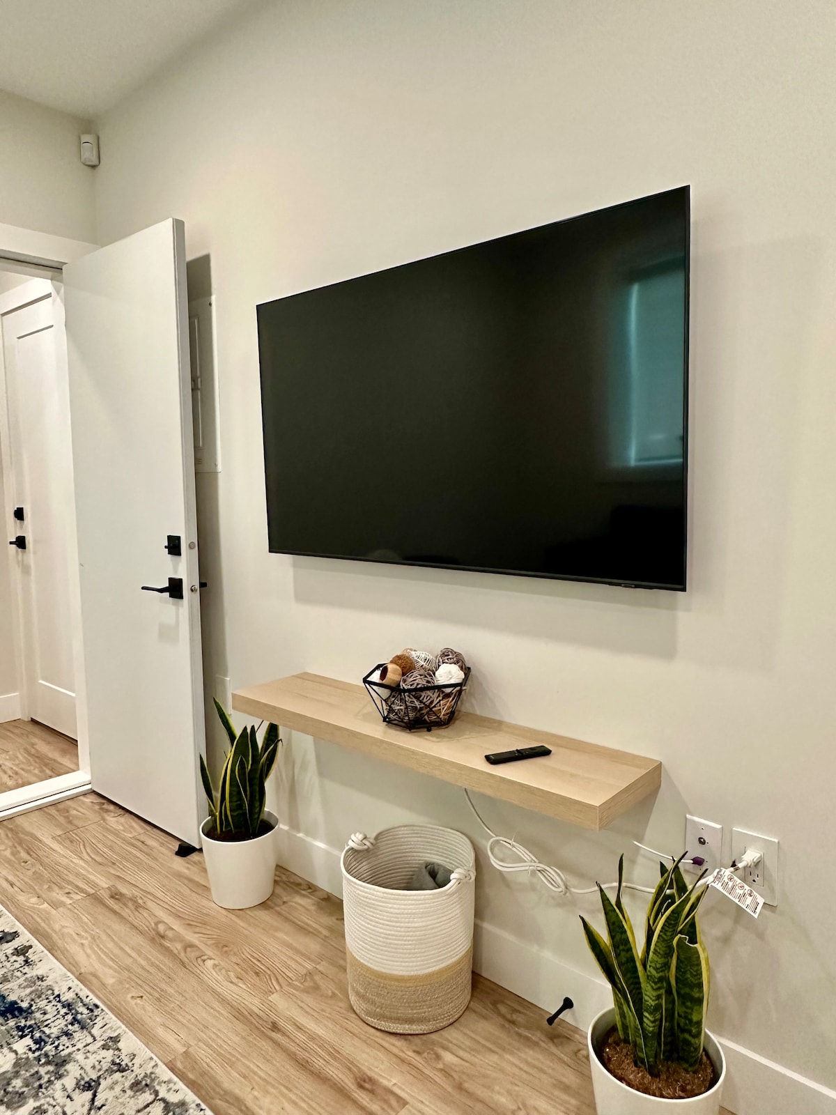 A minimalistic wall-mounted television is positioned over a wooden shelf, which features a decorative basket. Below, two potted plants are placed on the floor, one in a white woven planter and the other in a simple container. A white door in the background is slightly ajar.