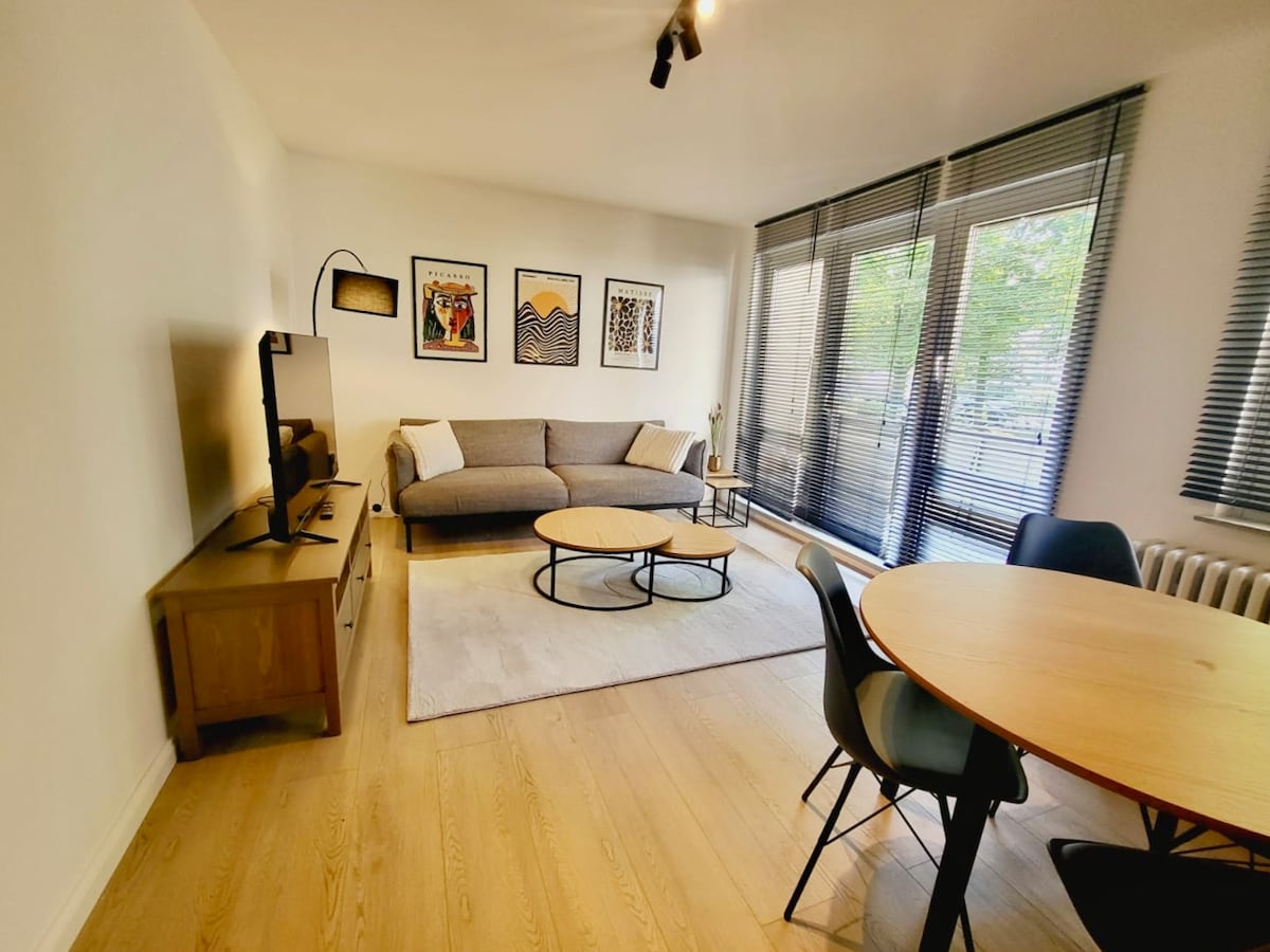 The living area features a comfortable gray sofa positioned across from a Smart TV. A round coffee table sits atop a soft rug, and a dining table with chairs is present nearby. Large windows offer natural light, partially covered by blinds.