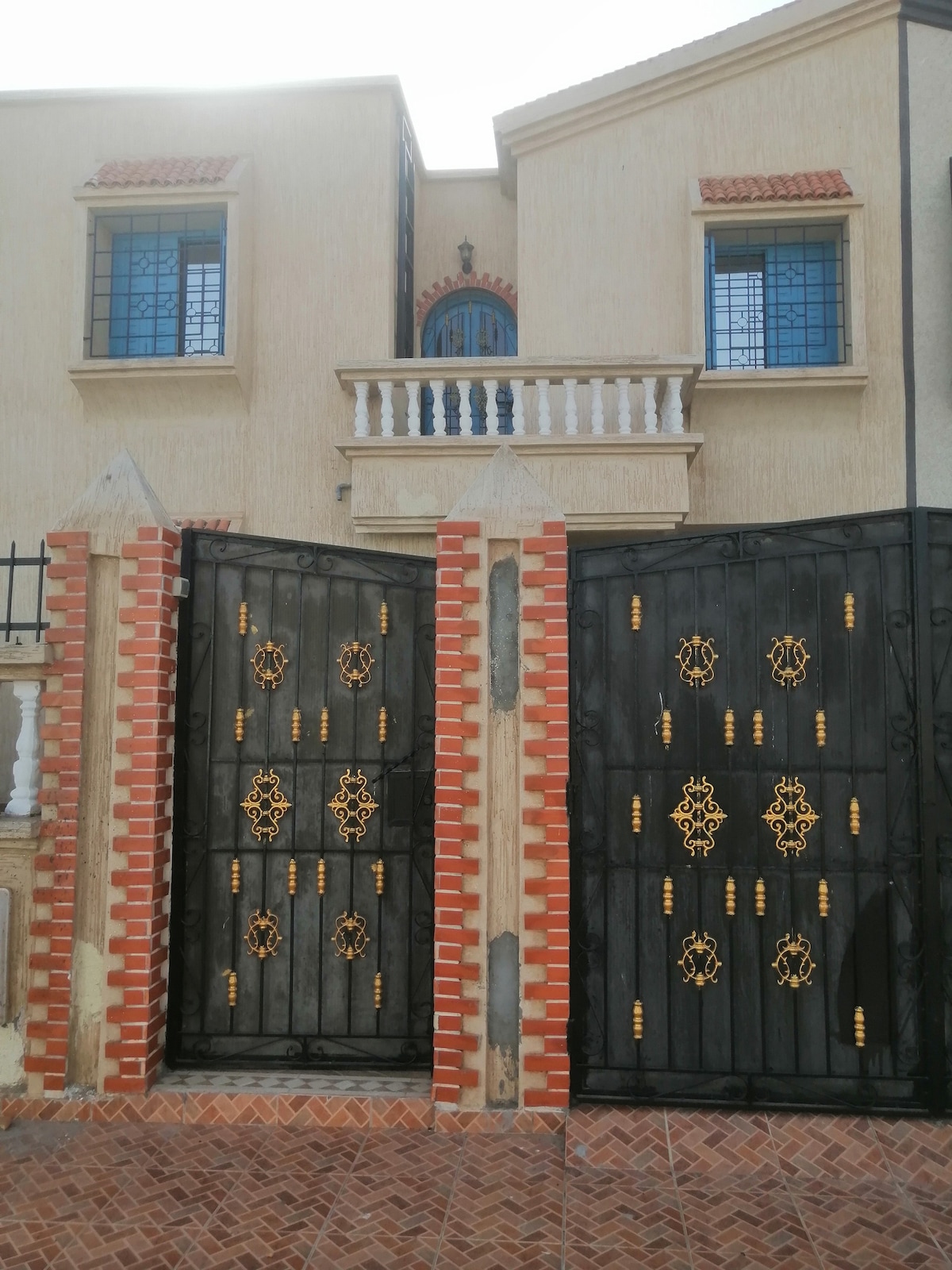 A decorative black iron gate with gold accents is framed by a contrasting brick wall. The entrance leads to a two-story building featuring arched windows and a balcony above. The warm sunlight highlights the entrance, adding to the welcoming appearance of the property.