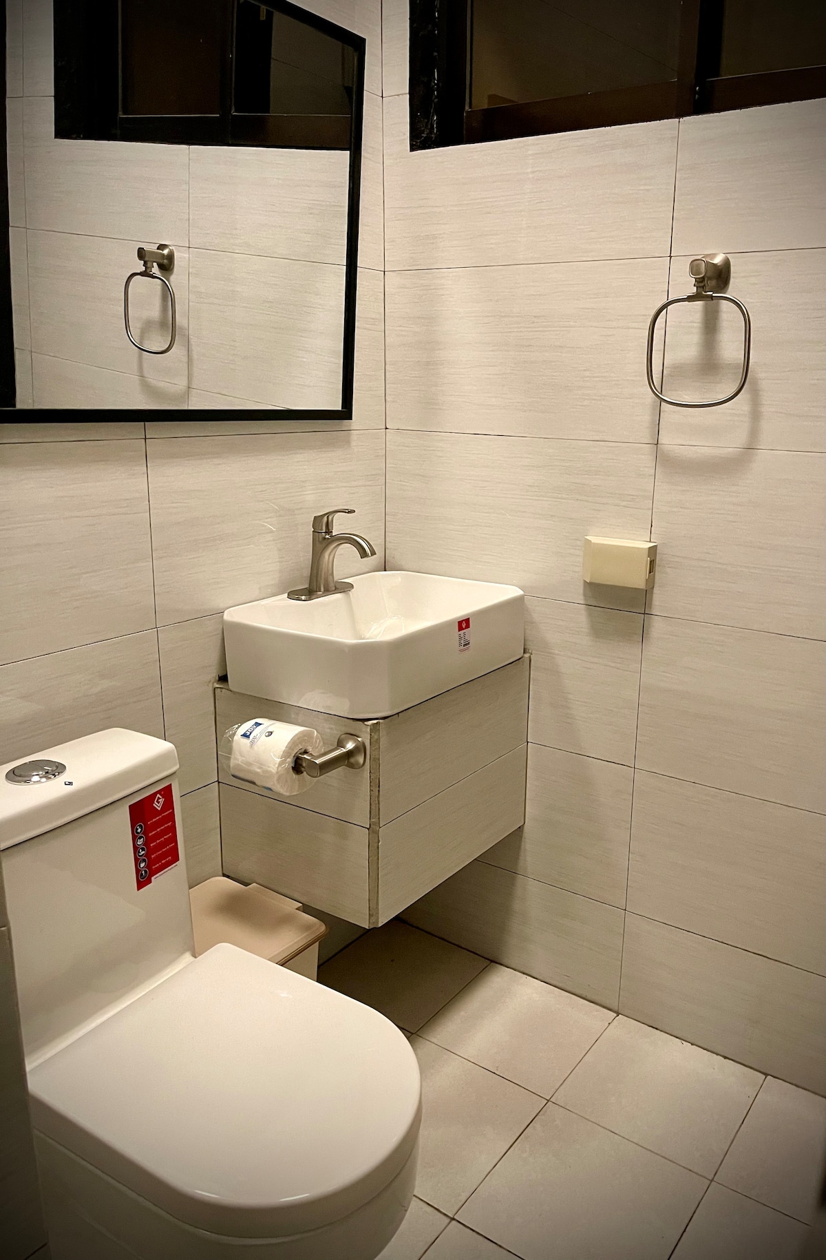 A modern bathroom features a sleek wall-mounted sink paired with a stylish faucet and a wall-mounted mirror. The toilet is positioned beside the sink, and chrome towel holders are mounted on the walls. Soft overhead lighting enhances the bright tiled space.