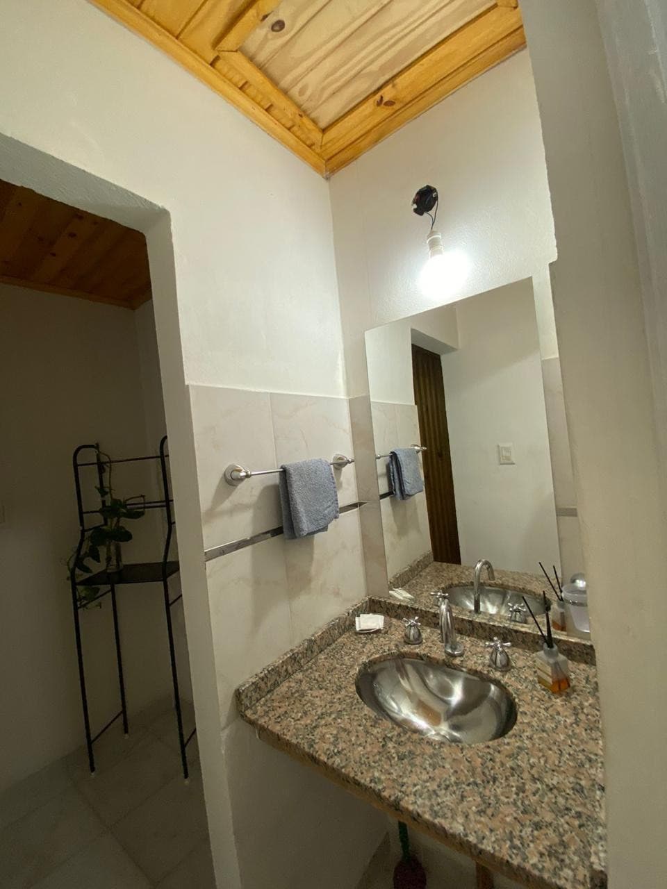 A clean bathroom is equipped with a granite countertop featuring a sink and chrome fixtures. Two neatly arranged towels hang on a towel bar. Natural light is provided by a small overhead light fixture, and a small shelving unit stands nearby, enhancing the functionality of the space.