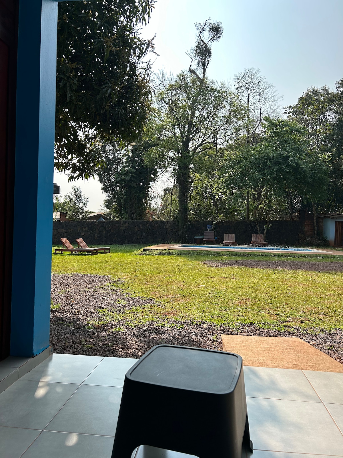 A green grassy area is viewed from a patio, framed by trees and shrubs. Sunlight illuminates the space, where lounge chairs and a fire pit are positioned in the distance. The ground is a mix of gravel and grass, providing a natural setting.