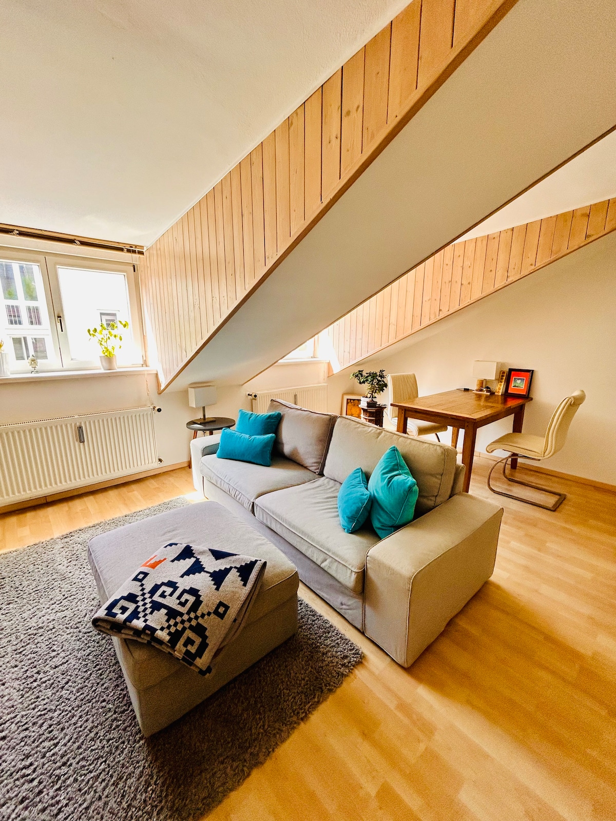 A light-filled living area is showcased with a spacious sofa adorned with decorative cushions. A cozy throw is draped over an ottoman. A wooden dining table with chairs is visible in the background, complemented by sloped wooden ceilings and large windows allowing natural light to enter.