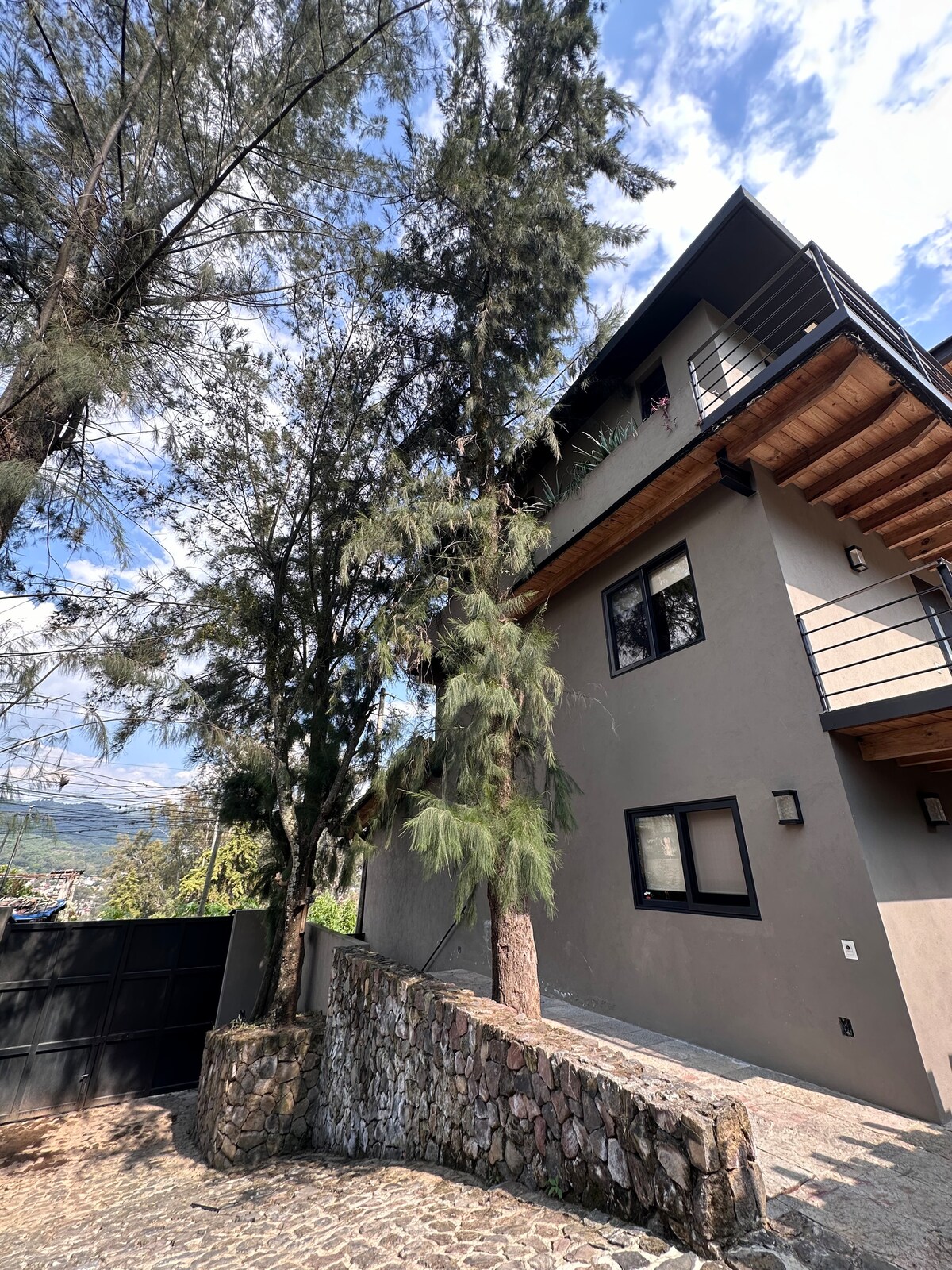 The exterior of the building showcases a modern design set against a backdrop of lush trees. The structure includes large windows, allowing for natural light, while a stone wall runs along the front, complementing the landscape.