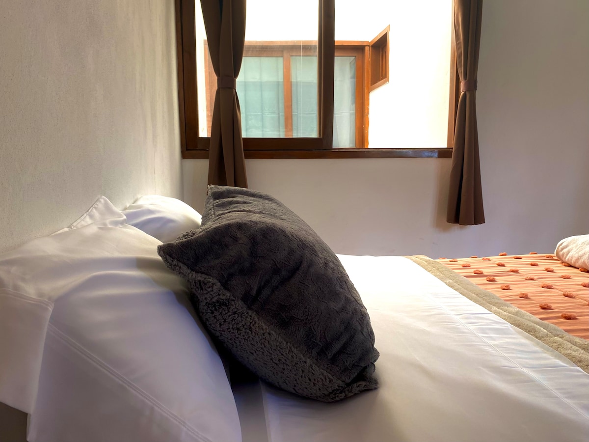 A spacious bed is dressed in crisp white linens, accompanied by a plush gray pillow. Natural light filters through a nearby window, which is framed by brown curtains, illuminating the cozy sleeping area.