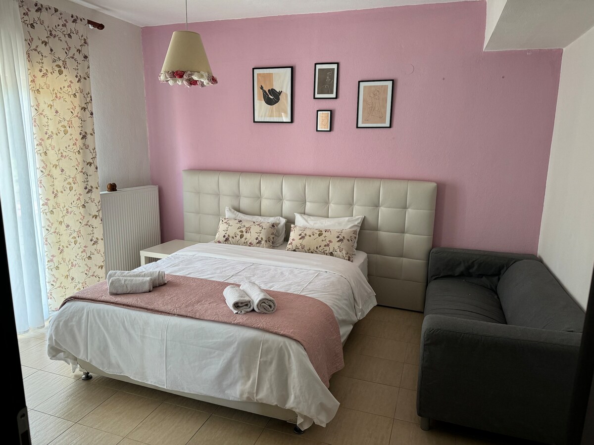 A light-filled bedroom features a spacious bed arranged with decorative pillows. A cozy sofa is positioned nearby, complemented by wall art and framed pictures. Natural light enters through a sheer curtain, enhancing the calming pink walls, while a floral accent adds a gentle touch.