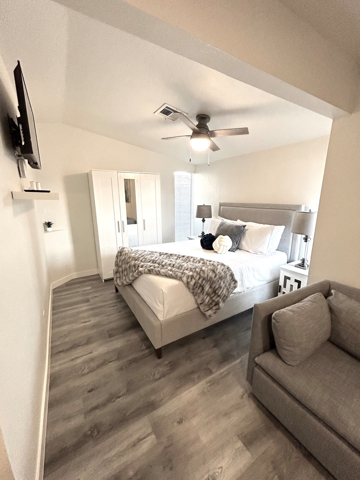A bright bedroom is furnished with a spacious bed draped in crisp linens. A light gray throw blanket is neatly placed at the foot of the bed. A cozy sofa rests in one corner, while a sleek wardrobe and wall-mounted TV enhance the modern decor.