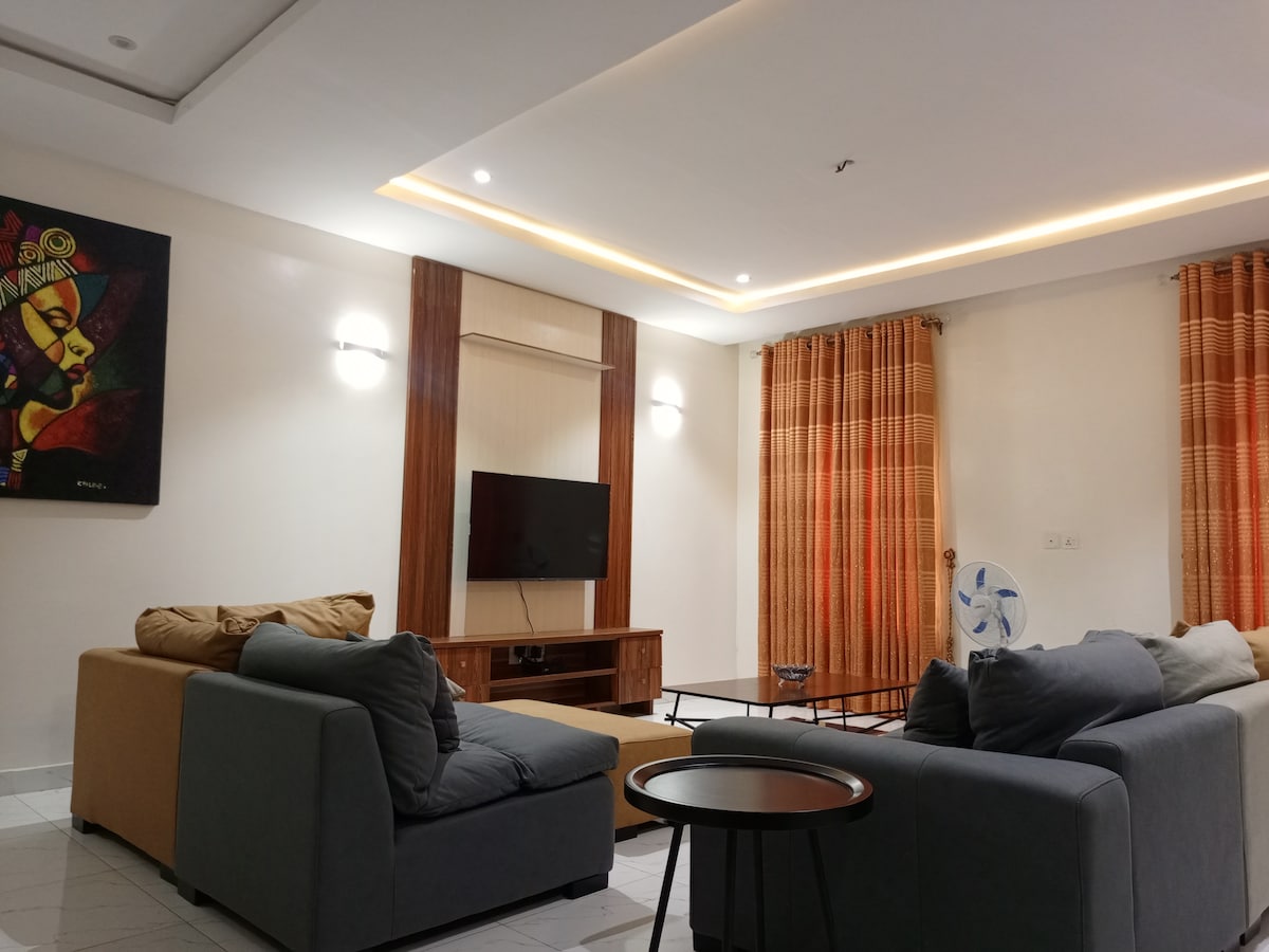 A spacious living room features two comfortable sofas, a coffee table, and a television mounted on the wall. Warm-toned curtains frame the windows, allowing soft light to fill the space. Decorative wall art adds a touch of character to the inviting environment.