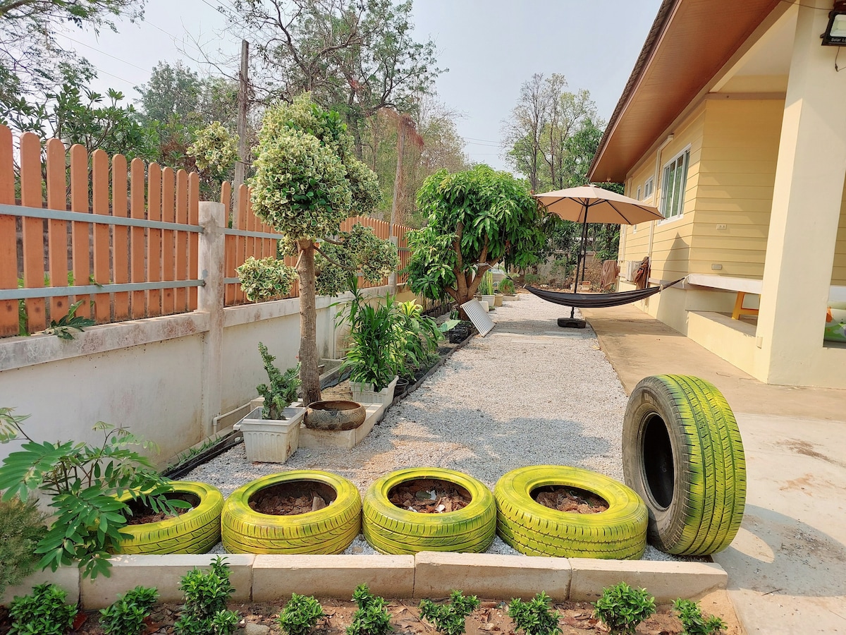 A garden area features a row of vibrant yellow tires, repurposed as planters. Lush greenery thrives nearby, while a gravel pathway leads to a shaded seating space equipped with a hanging hammock and an umbrella for relaxation. A well-manicured hedge lines the background.