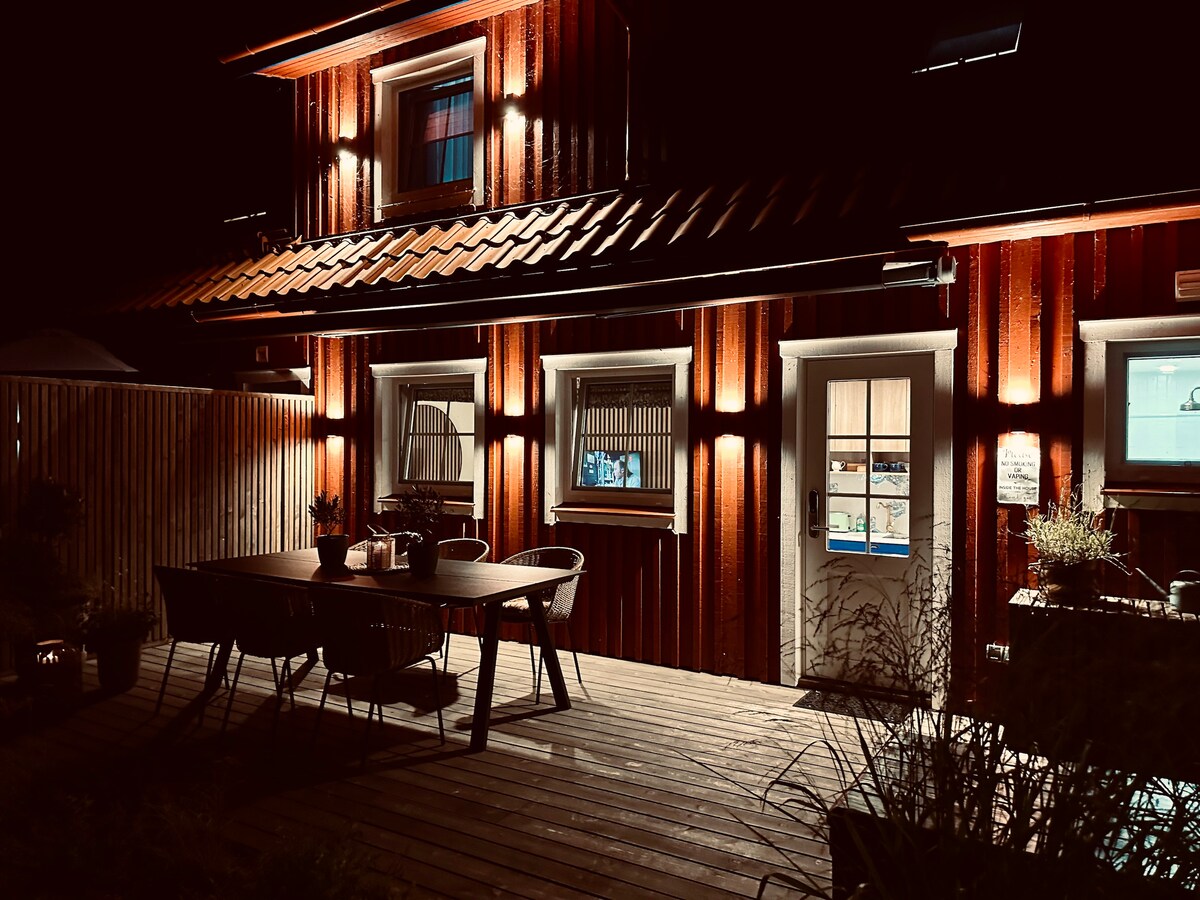 The exterior of the building is illuminated at night, highlighting the warm wooden facade. A spacious wooden deck features a table surrounded by chairs, creating an inviting outdoor dining area. Soft lighting enhances the entrance, complementing the rustic charm.