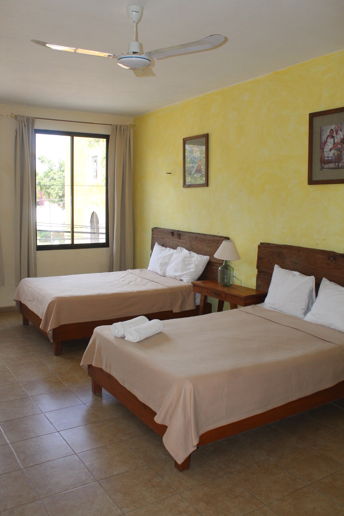 A brightly lit room features two spacious beds with cream-colored bedspreads. Natural light streams in through large windows adorned with light curtains. A bedside table holds a lamp, while framed artwork decorates the yellow walls, creating a comfortable and inviting atmosphere.