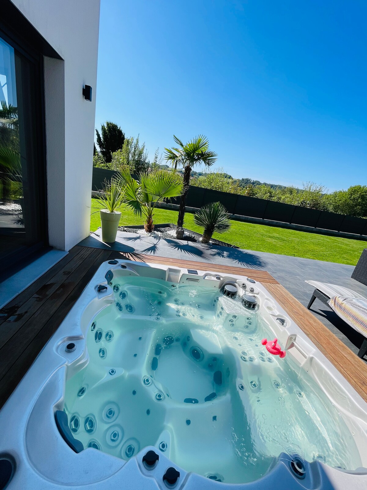 A private therapeutic spa is shown, surrounded by lush greenery and a clear blue sky. A wooden deck extends beside the spa, which features various jets and seating areas. Tall palm trees and other plants are visible in the background.