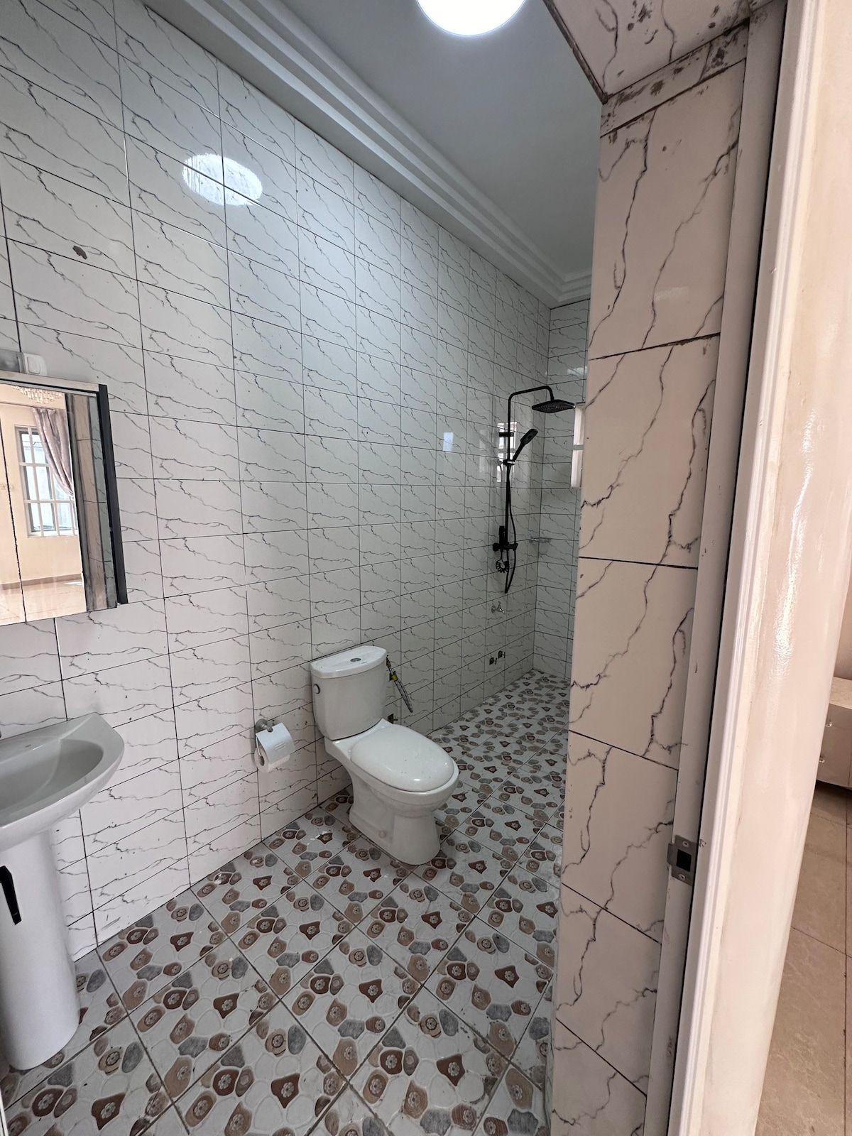 An expansive bathroom is presented, featuring a modern toilet and a spacious walk-in shower. The walls are tiled with a subtle marble pattern, while the floor exhibits a decorative design. A mirror above the sink enhances the sense of space and brightness.