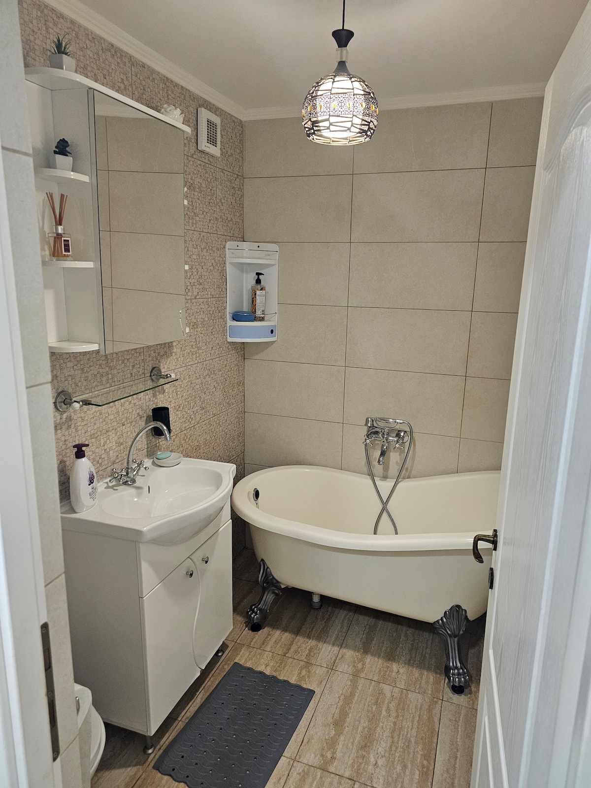 A compact bathroom showcases a freestanding bathtub with elegant claw feet. A modern sink is placed beside a wall-mounted mirror. Neutral tile surrounds the space, complemented by a decorative hanging light fixture. Shelves above the sink hold toiletries, maintaining a clean and organized look.