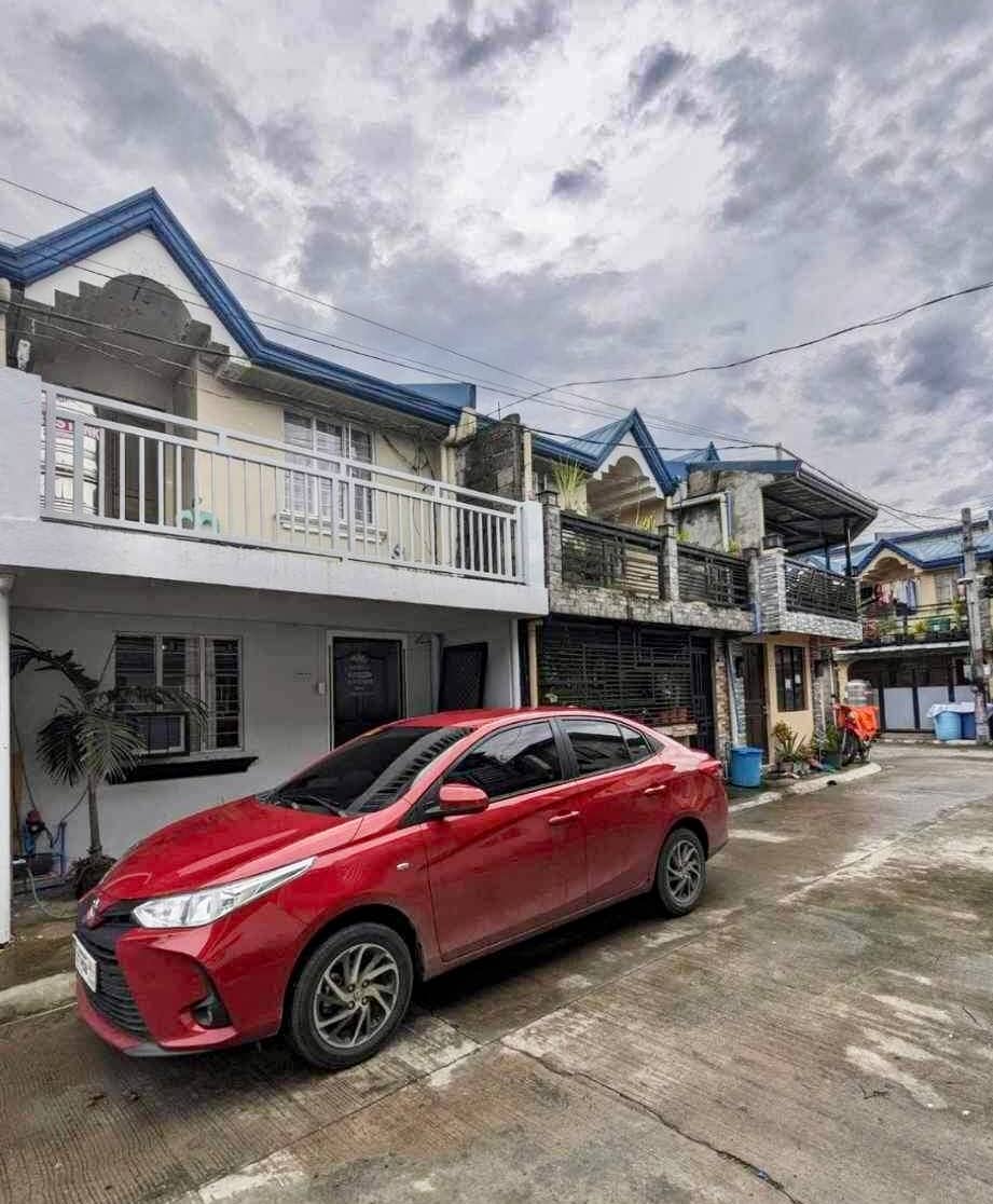 A red car is parked in front of a two-story house, featuring a balcony on the upper level. The house is surrounded by a well-maintained courtyard, while a cloudy sky looms above, creating a serene atmosphere. Nearby, there are signs of residential activity.