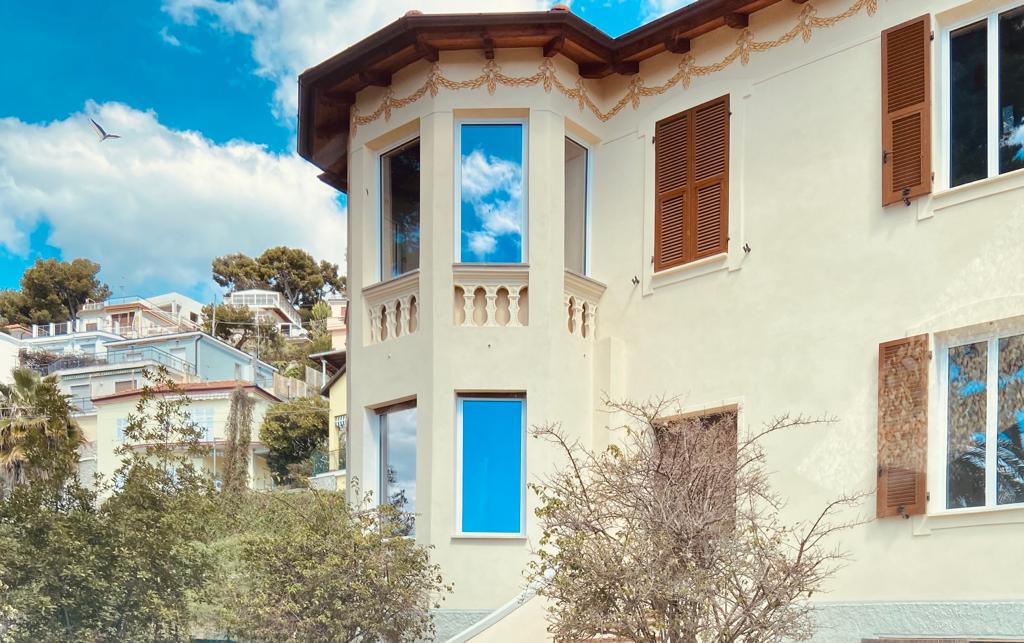 The exterior of the villa is displayed, featuring a unique architectural design with large windows and decorative details. The light-colored walls contrast with dark shutters, while surrounding greenery adds a natural element. A clear blue sky and distant hillside provide a scenic backdrop.