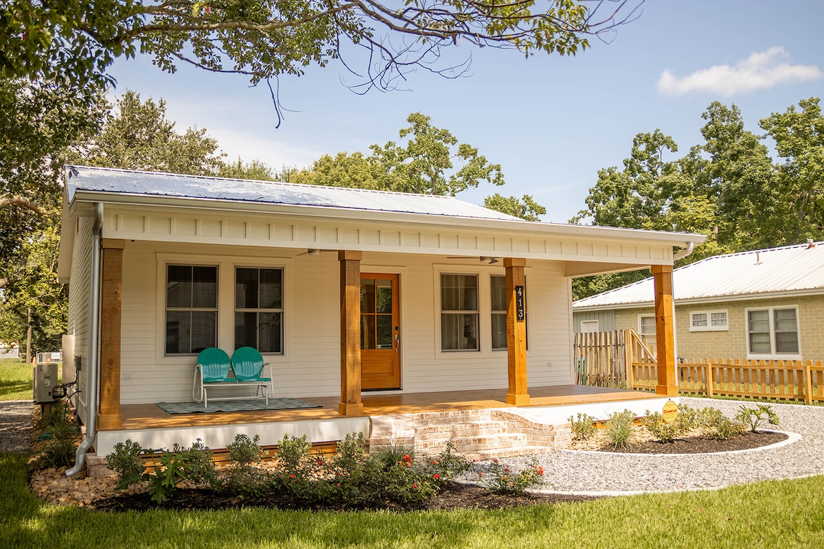 A charming single-story house with a welcoming front porch featuring two teal chairs. The home is surrounded by a well-kept lawn and landscaped flower beds, complemented by green trees in the background under a clear blue sky.