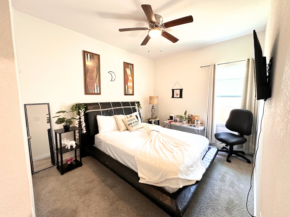 A bedroom with a spacious bed placed against a black headboard. Two framed artworks hang above the bed, and a ceiling fan provides airflow. A mirror and a workstation are included, with natural light streaming through a window covered by light curtains.
