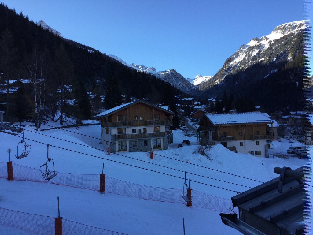 The image shows a winter landscape with snow-covered ground and mountains in the background. Two chalet-style buildings are visible, surrounded by trees. Ski lifts are seen in the foreground, providing access to the slopes. A clear blue sky complements the scenic view.