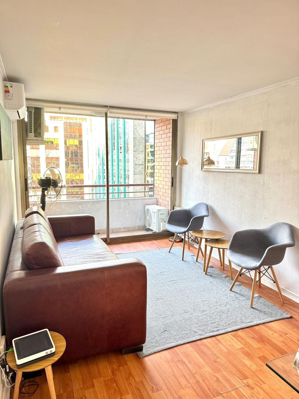 A living area features a brown leather sofa paired with two gray chairs and a small round table. A large window allows natural light to fill the space, while a wall-mounted mirror reflects the surroundings. Wood flooring extends throughout the area.