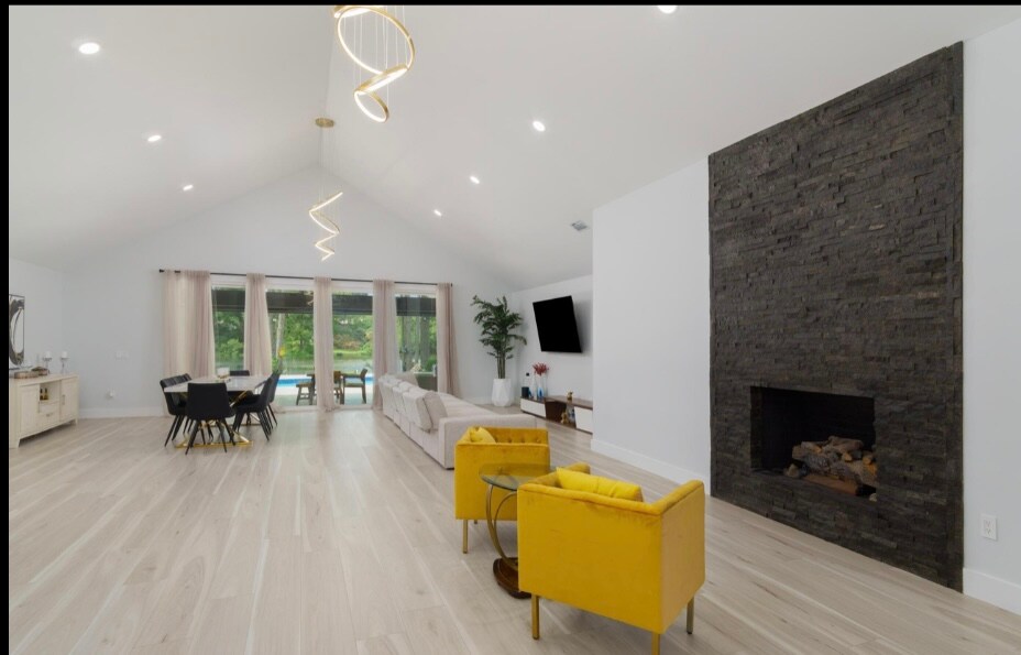 A spacious living area features high ceilings and large windows offering views of the outdoor space. A sleek stone fireplace is positioned against a pale wall. Yellow armchairs add a pop of color, while a dining table for four is set nearby, enhancing the open layout.